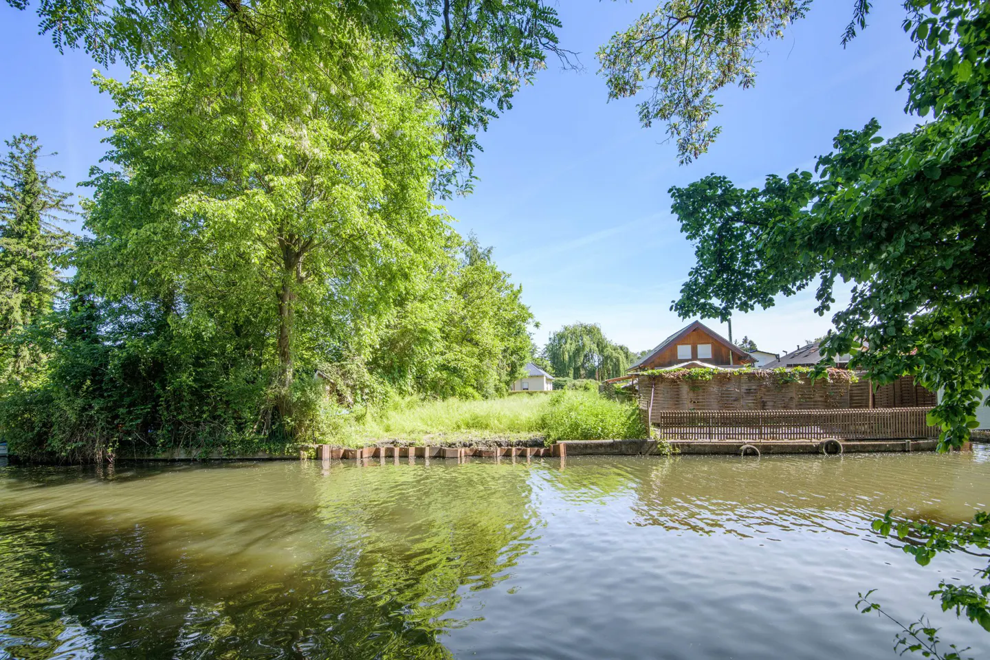 A calm river flows past a grassy bank lined with lush green trees and a wooden fence under a clear blue sky.