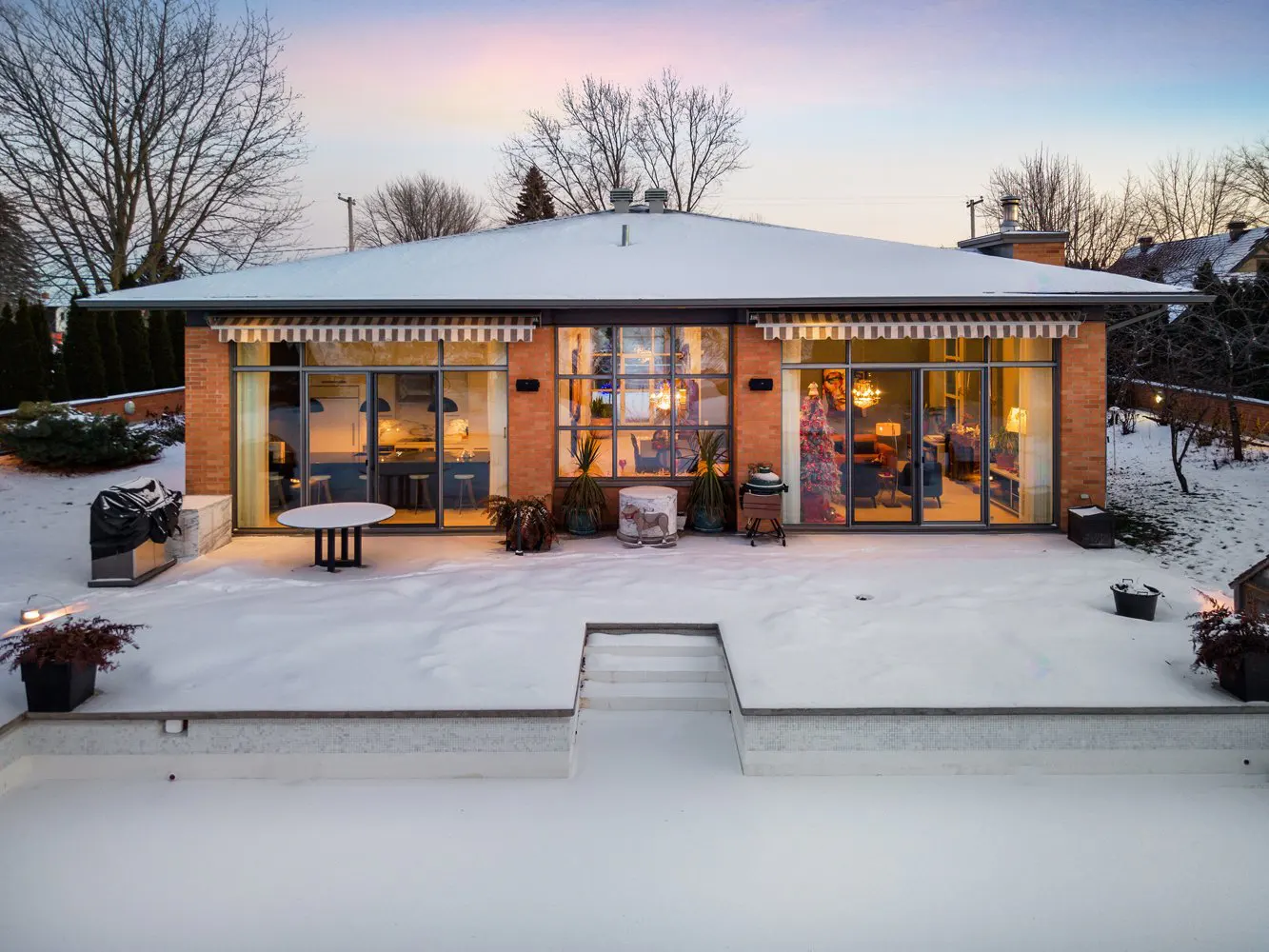 Back view of a brick house with large windows and a snow-covered patio at dusk. Interior lights are on.
