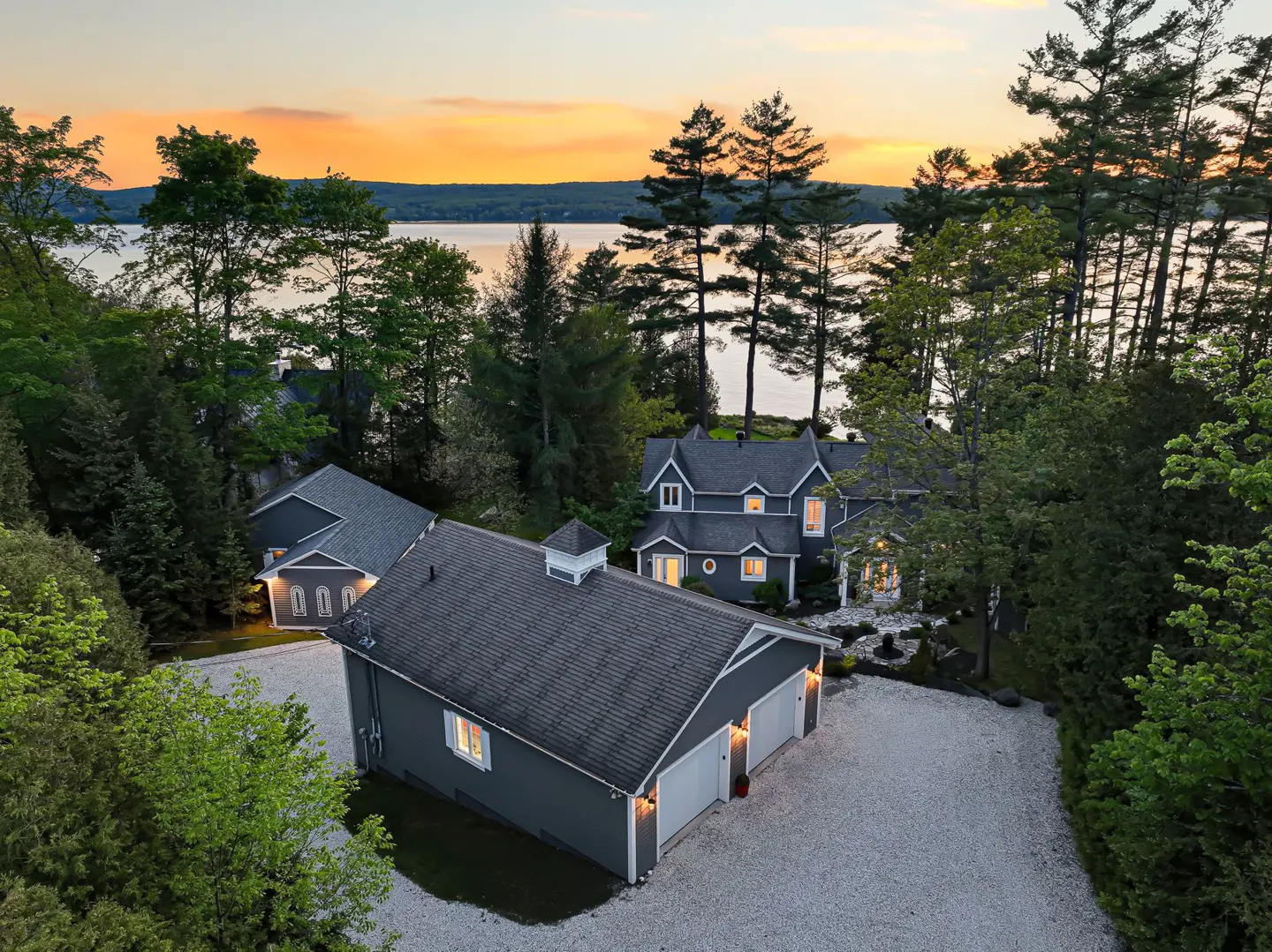 Aerial view of a grey lakeside home and detached garage surrounded by lush trees at sunset with a glowing orange sky.