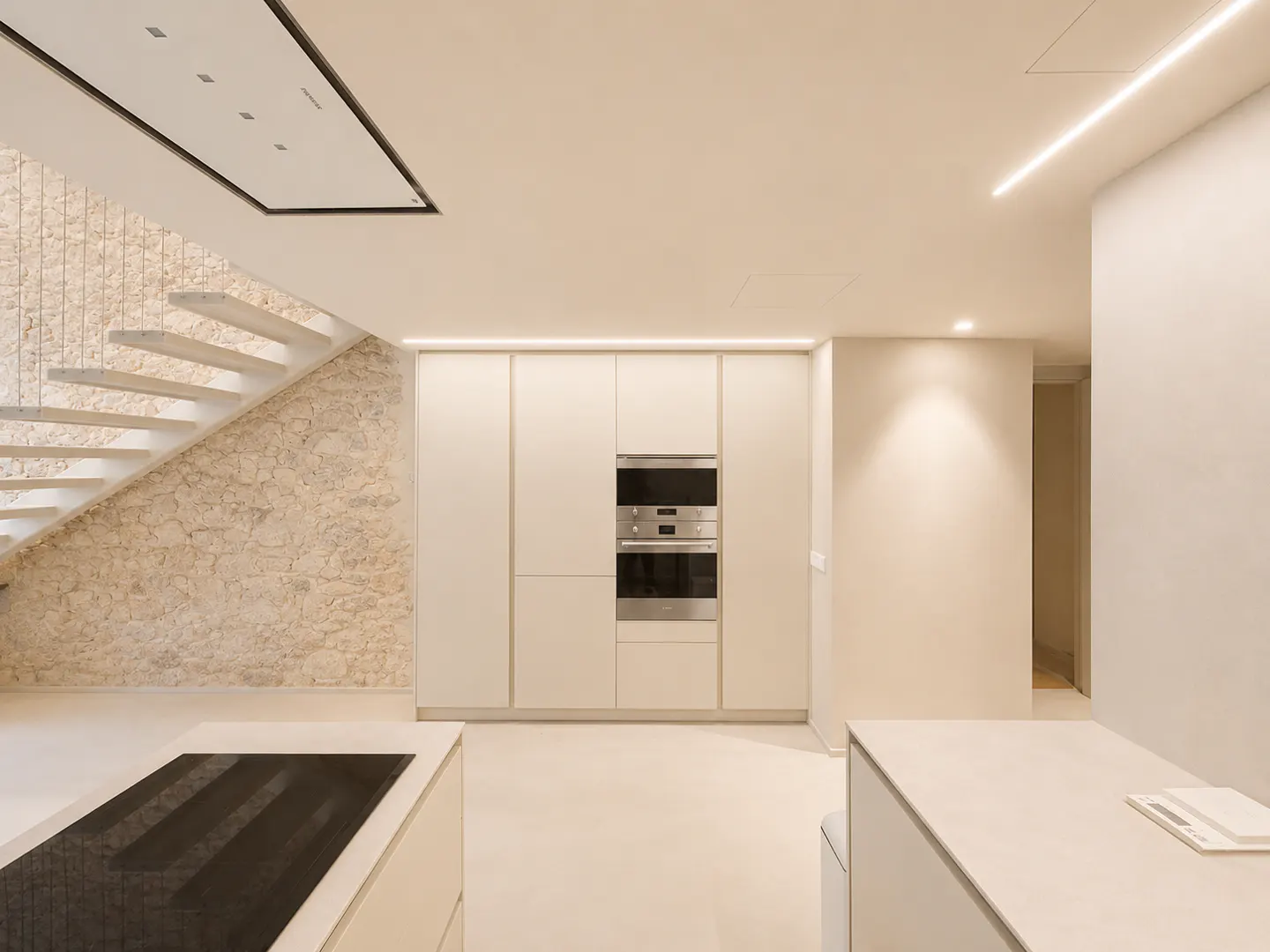 Minimalist kitchen with cream cabinets, stainless steel ovens, and a stone wall staircase.