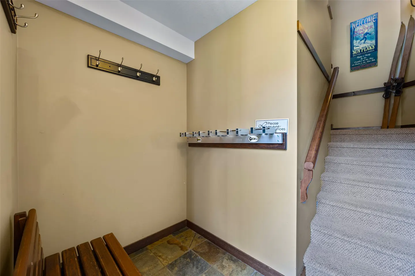 Entryway with coat hooks, shoe rack, and bench. Stairs with skis and a "Welcome Winter" sign are visible. The walls are beige.