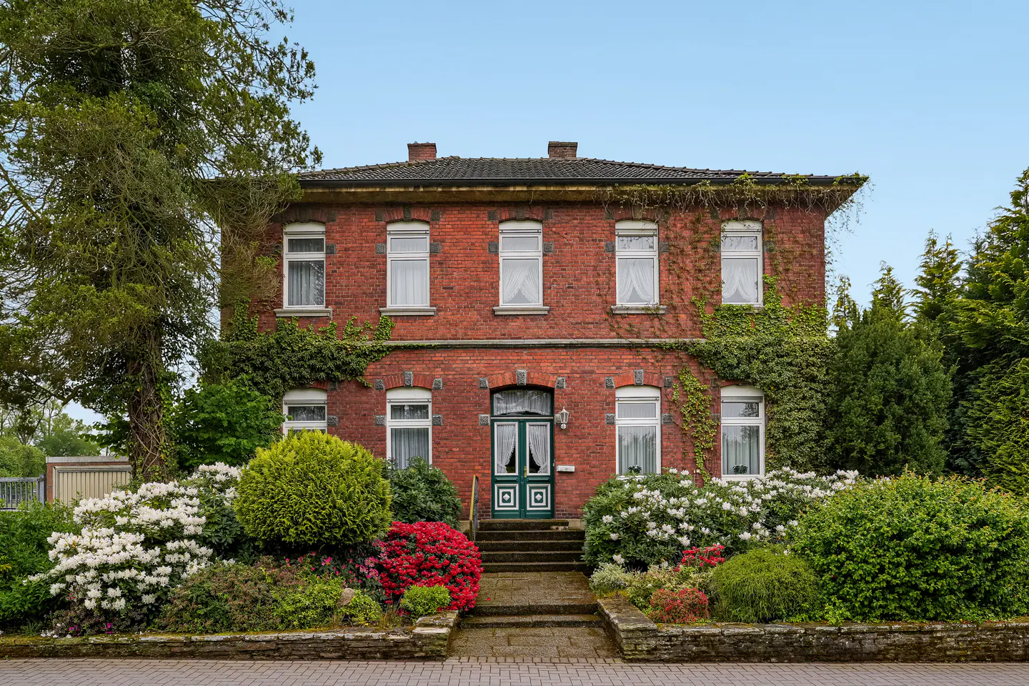 Two-story red brick house with white-framed windows, green door, and lush garden with colorful flowers.