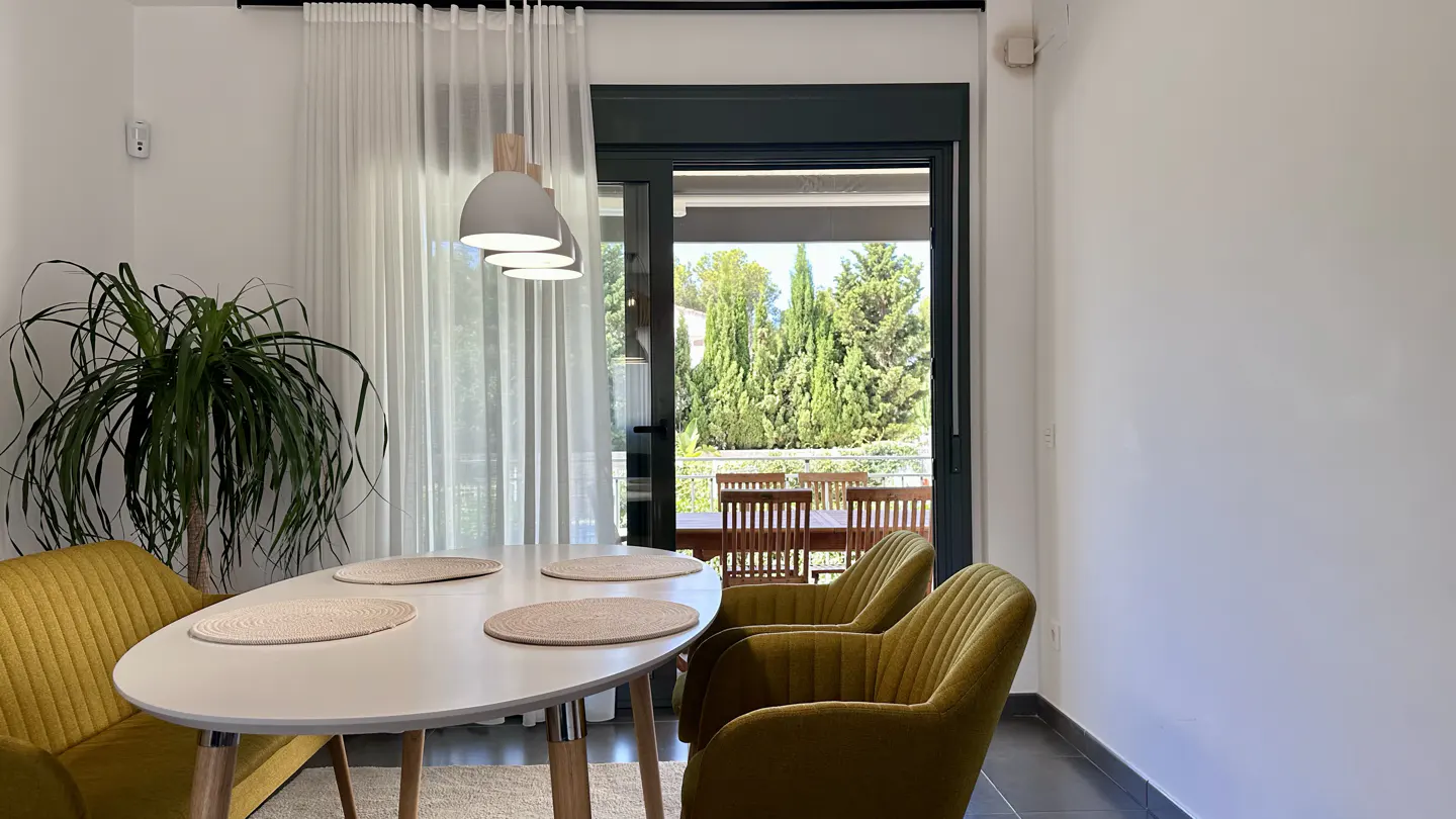 A bright dining room with a white table, olive green chairs, and a view of a green garden through a sliding glass door.