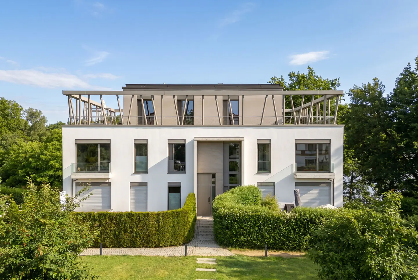 Modern white three-story apartment building with a unique rooftop pergola, surrounded by lush green hedges and trees.