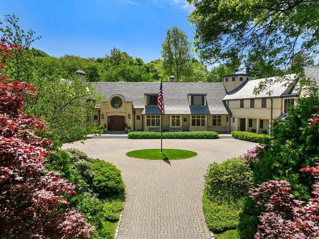 Beige mansion with a circular driveway, an American flag, and lush green landscaping under a blue sky.