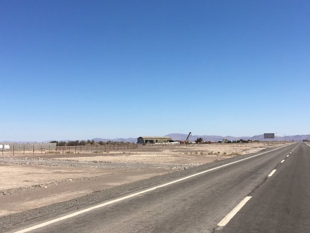 A long asphalt road stretches into the distance under a clear blue sky. To the left, a fenced, vacant lot with a building and construction equipment.