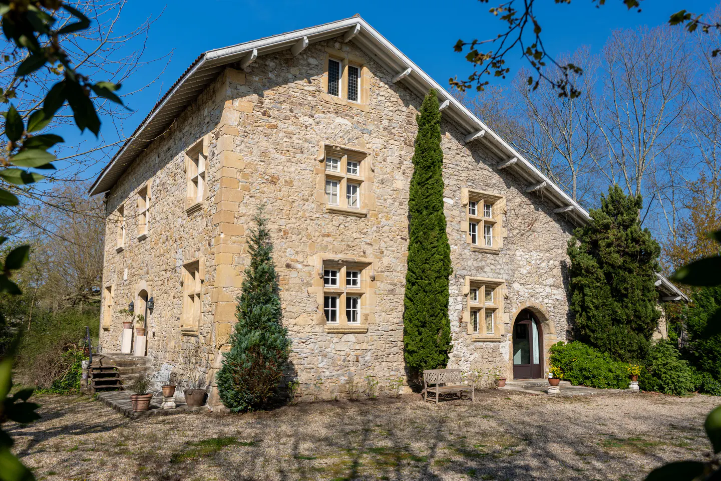 Stone house with white trim, multiple windows, and a bench in front. Trees surround the house.