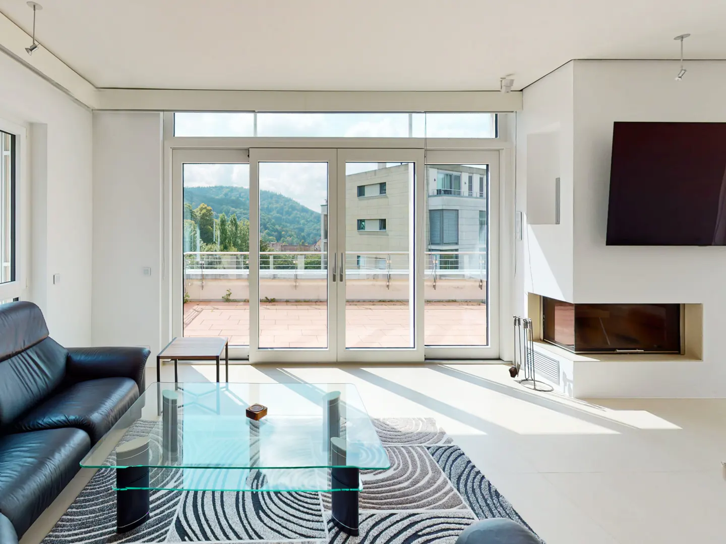 Bright living room with a black leather sofa, glass table on a patterned rug, and a fireplace with a TV above it. Sliding glass doors lead to a balcony with a view.
