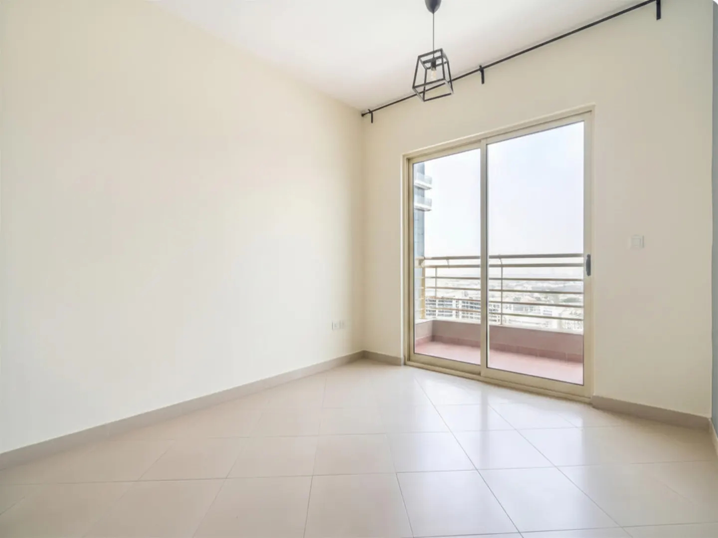 An empty room with beige walls and tile floor. A sliding glass door leads to a balcony with a city view. A modern light fixture hangs from the ceiling.