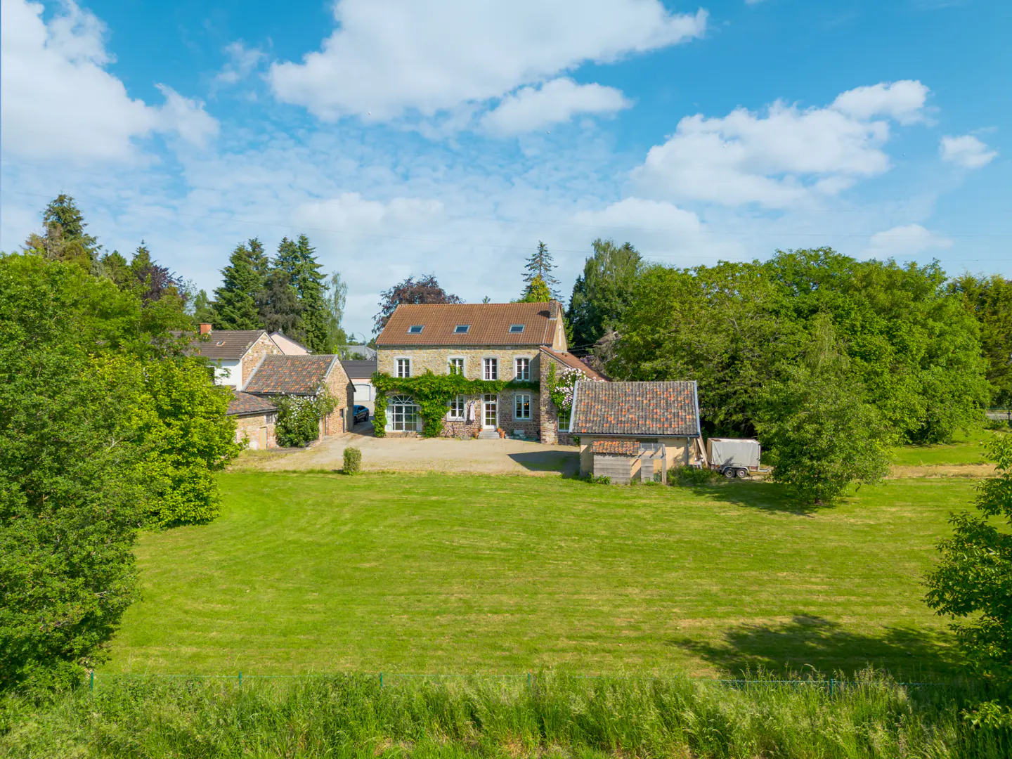 A stone farmhouse with a tiled roof and climbing vines, set in a large green field surrounded by trees under a blue sky.