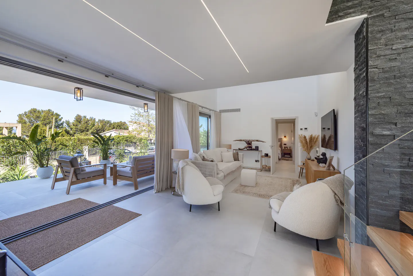 Bright, modern living room with white walls, light wood floors, and a stone accent wall. Sliding glass doors open to a patio with outdoor seating.