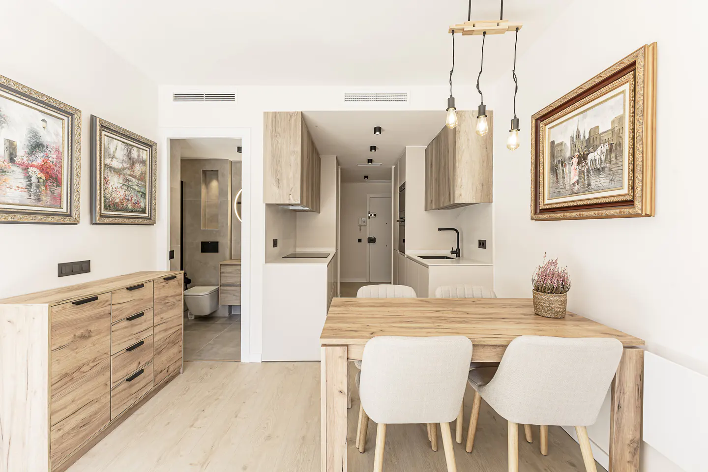 Bright apartment interior with wood dining table, chairs, and pendant lights. A kitchen and bathroom are visible in the background. Two framed paintings hang on the wall.