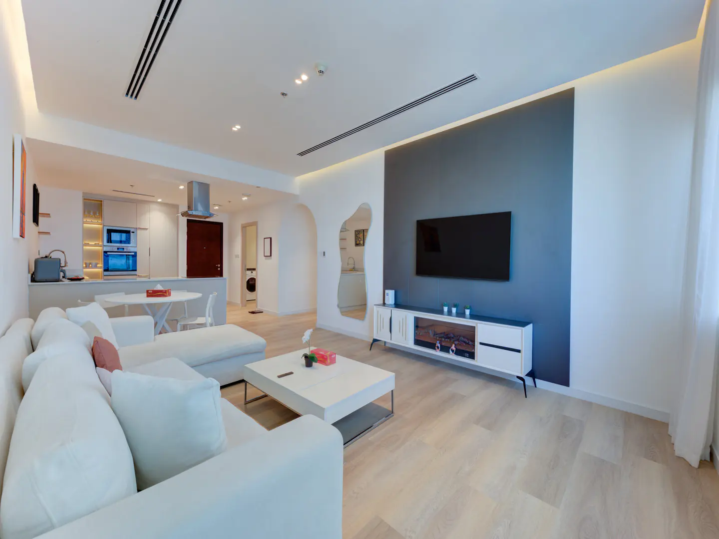 Bright, modern apartment with a white sectional sofa, TV, and open kitchen. Light wood floors and gray accent wall.