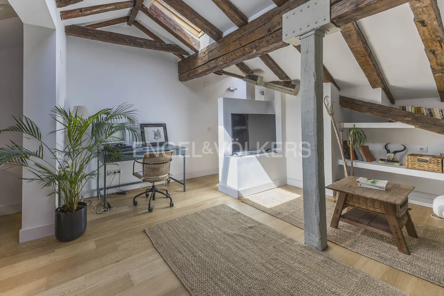 Attic room with exposed beams, skylight, and wooden floors. A desk, chair, and plant are on the left, and a TV and shelves are on the right.