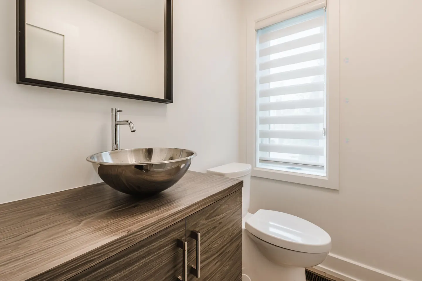 Modern bathroom with a stainless steel bowl sink, wood cabinet, black framed mirror, white toilet, and a window with blinds.