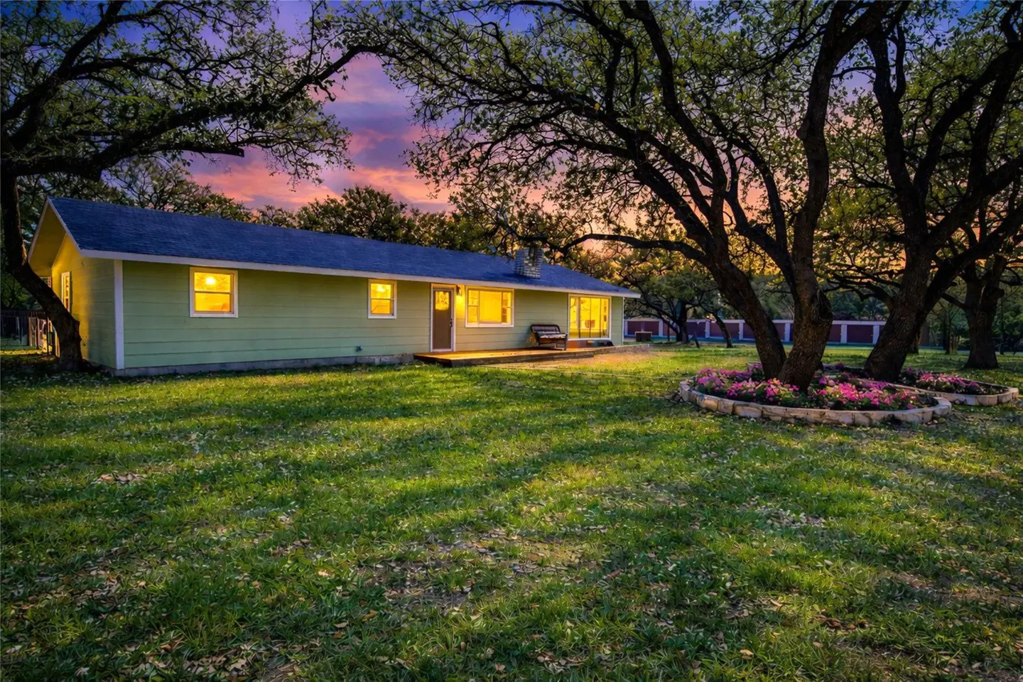 A single-story, light green house with a blue roof, surrounded by large trees and a green lawn at dusk.