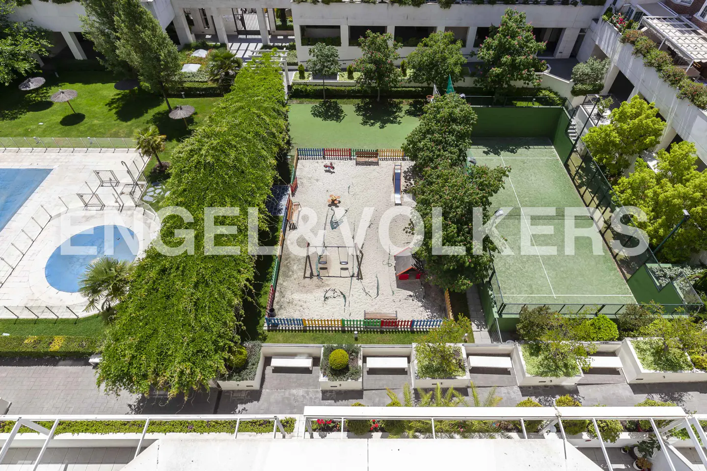 Aerial view of a community area with a pool, playground, tennis court, and green spaces. The area is surrounded by buildings.