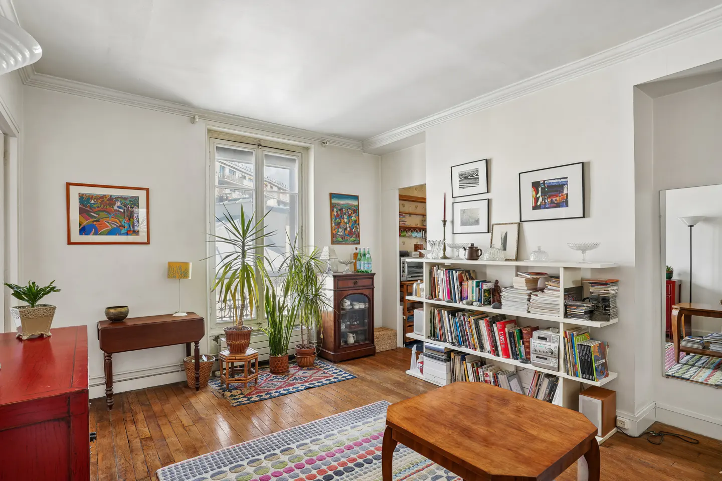 Bright living room with hardwood floors, white walls, and a large window. Plants, a bookshelf, and colorful rugs add character.