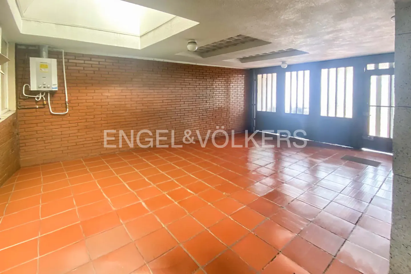 A spacious room with orange tile flooring, brick walls, and a water heater. Large, dark doors with windows are visible.