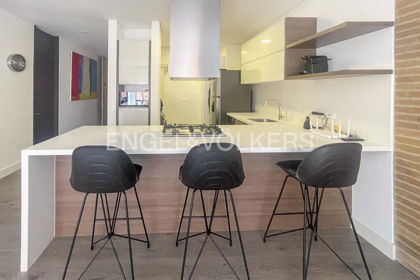 A modern kitchen with a white countertop island and three black bar stools. Stainless steel appliances and light wood accents are visible.