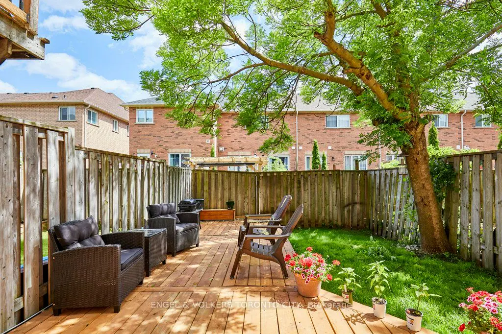 A wooden backyard deck with wicker patio chairs, potted flowers, and a green lawn enclosed by a tall wooden fence.