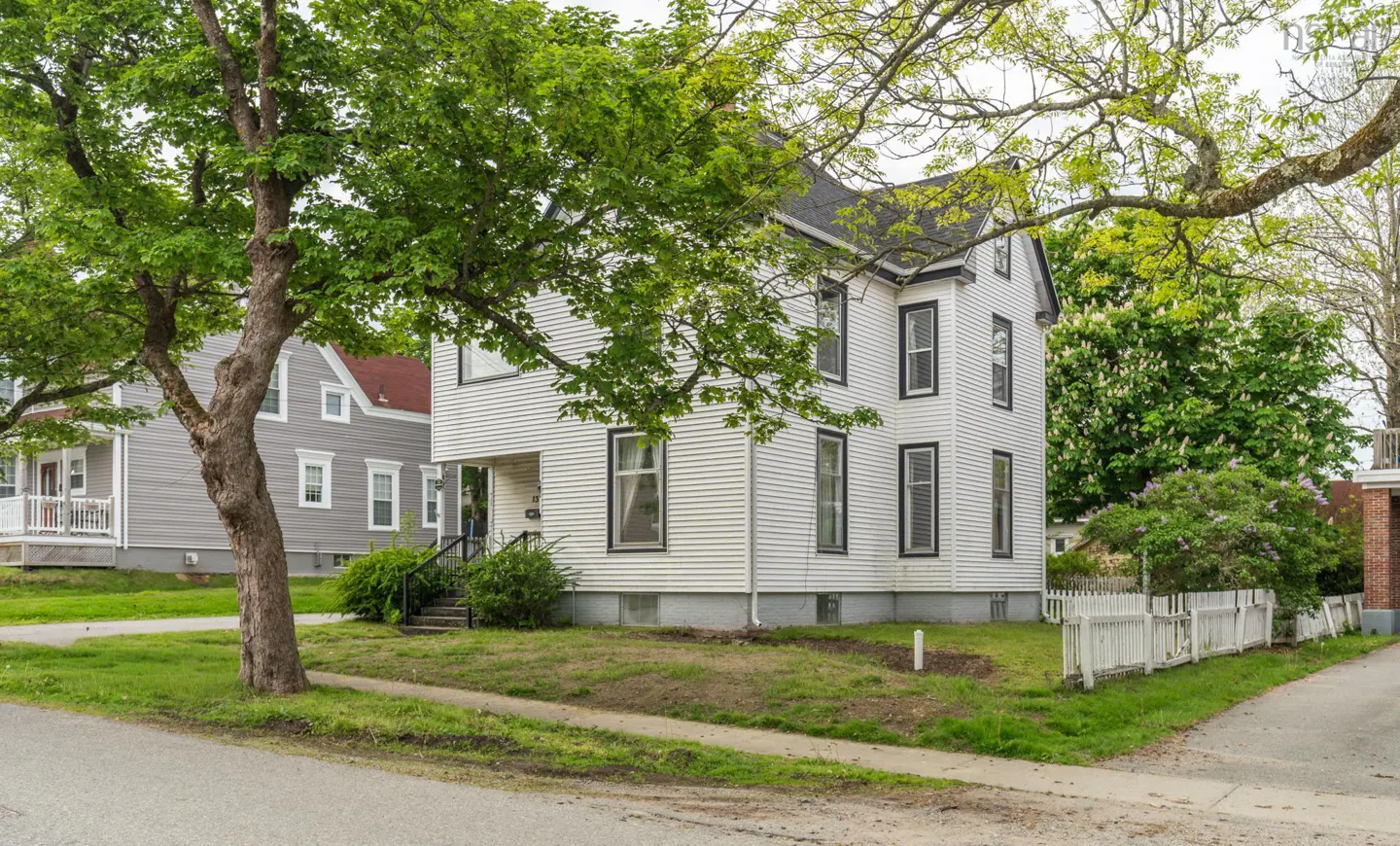 Two-story white house with black trim, surrounded by green trees and a white picket fence on a residential street.