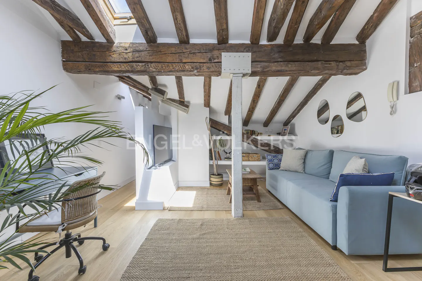 Bright living room with exposed wooden beams, light blue sofa, and jute rug. A desk and chair are on the left.