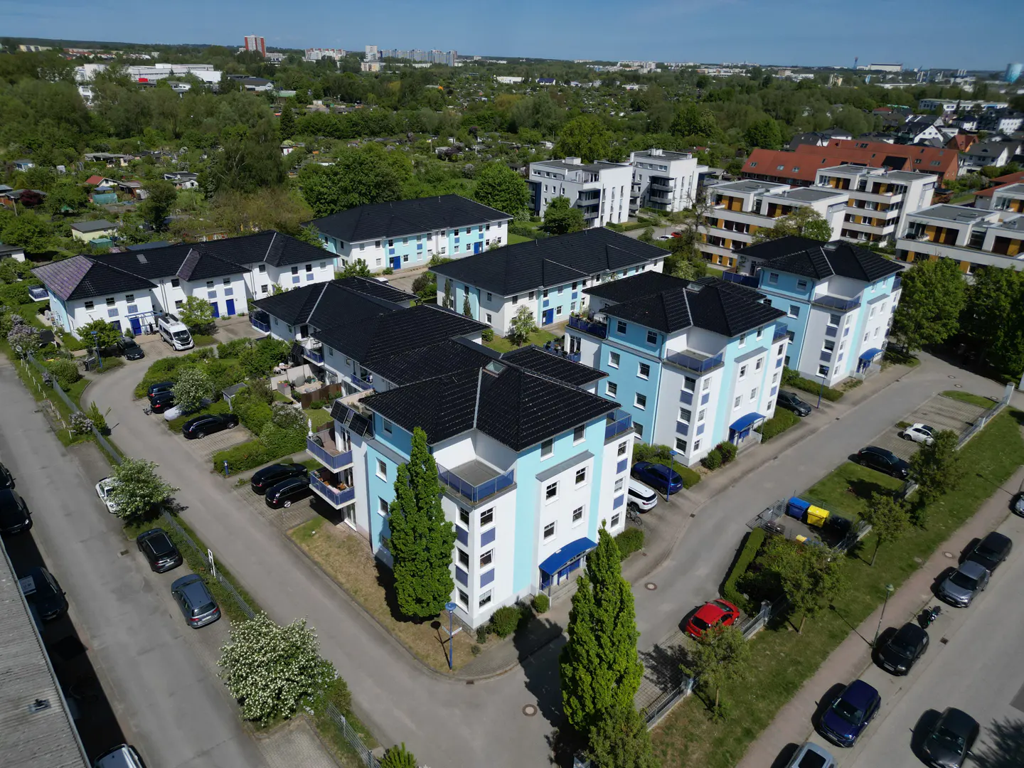 Aerial view of a residential complex with light blue and white buildings, black roofs, and surrounding parked cars.