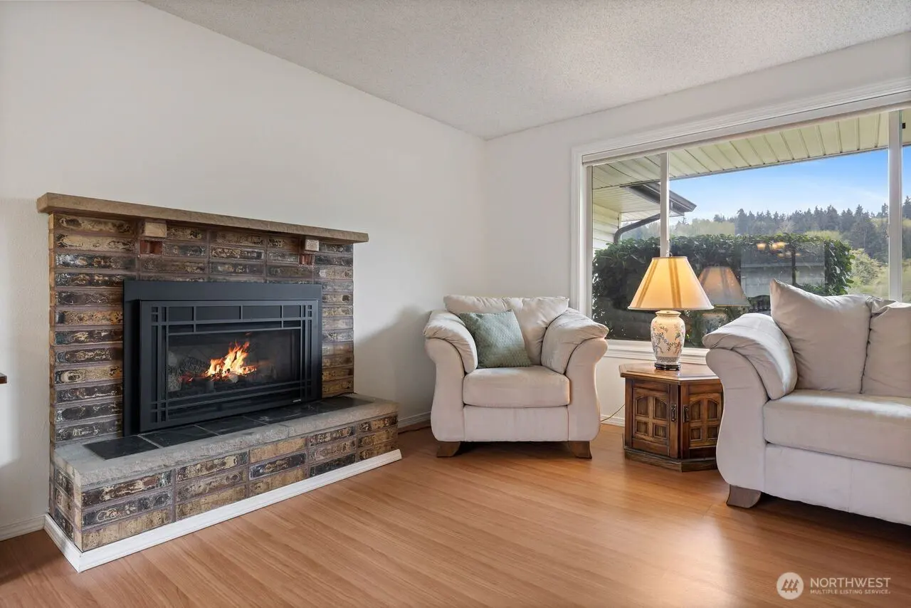 Living room with brick fireplace, light wood floors, and white sofa and armchair near a large window.