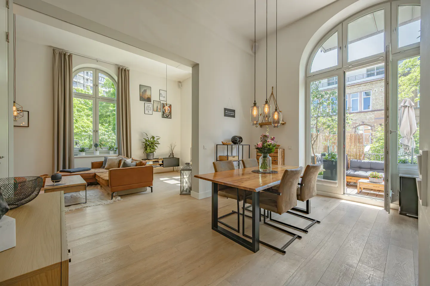 Open-concept living and dining area with wood floors, brown leather sofa, dining table with four chairs, and arched windows leading to a patio.