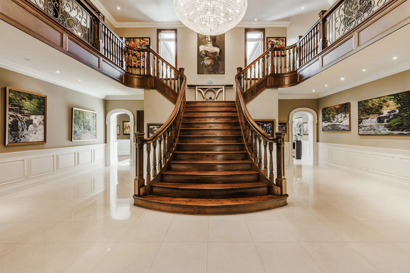 Grand foyer with a curved wooden staircase, chandelier, and art. Beige walls, white trim, and a shiny tile floor.