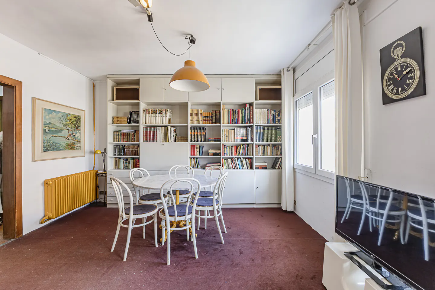 A room with a round table, white chairs, a bookshelf filled with books, and a painting on the wall.