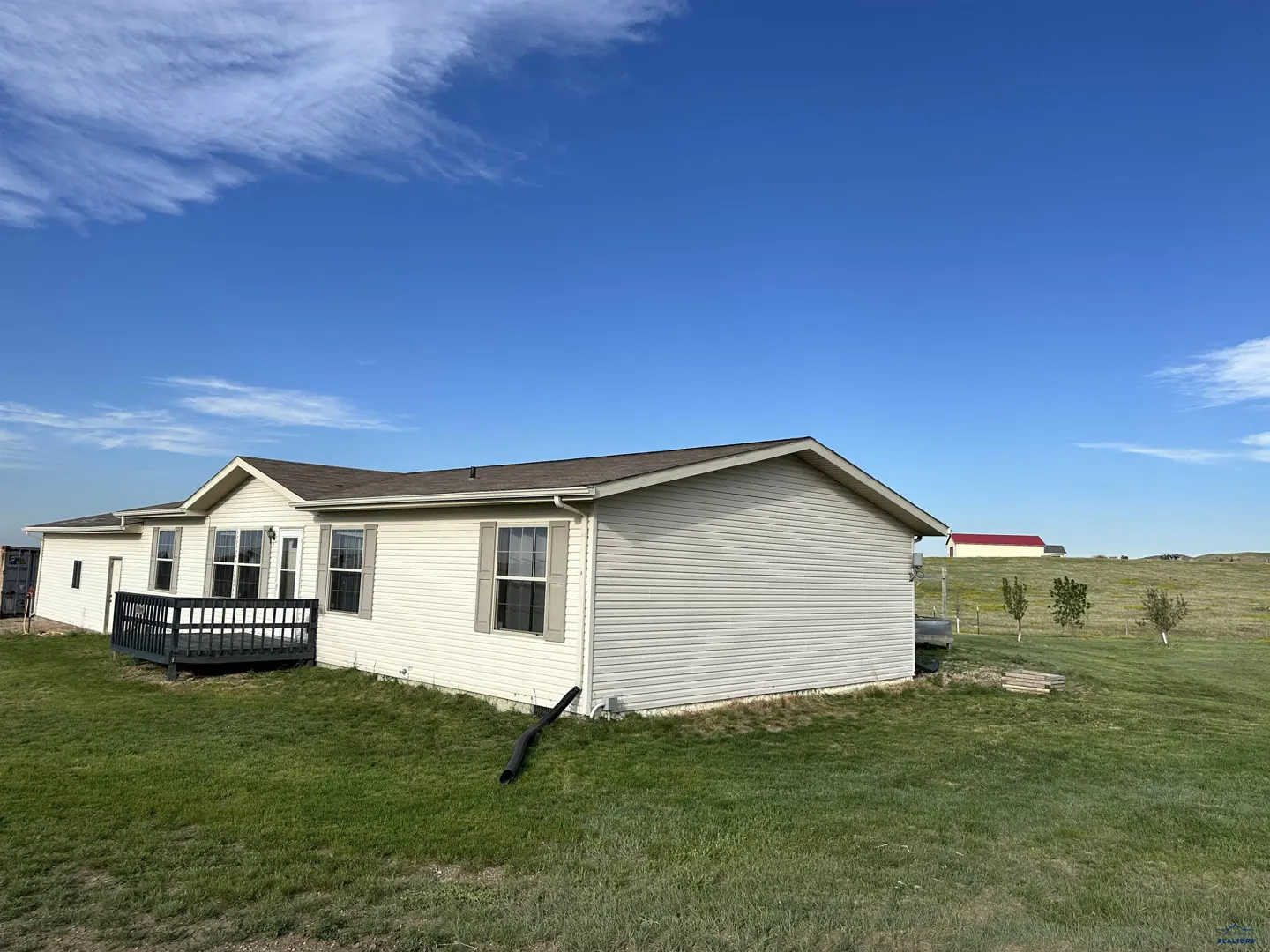 Single-story beige home with a dark deck, set in a grassy field under a clear blue sky with a distant red-roofed barn.
