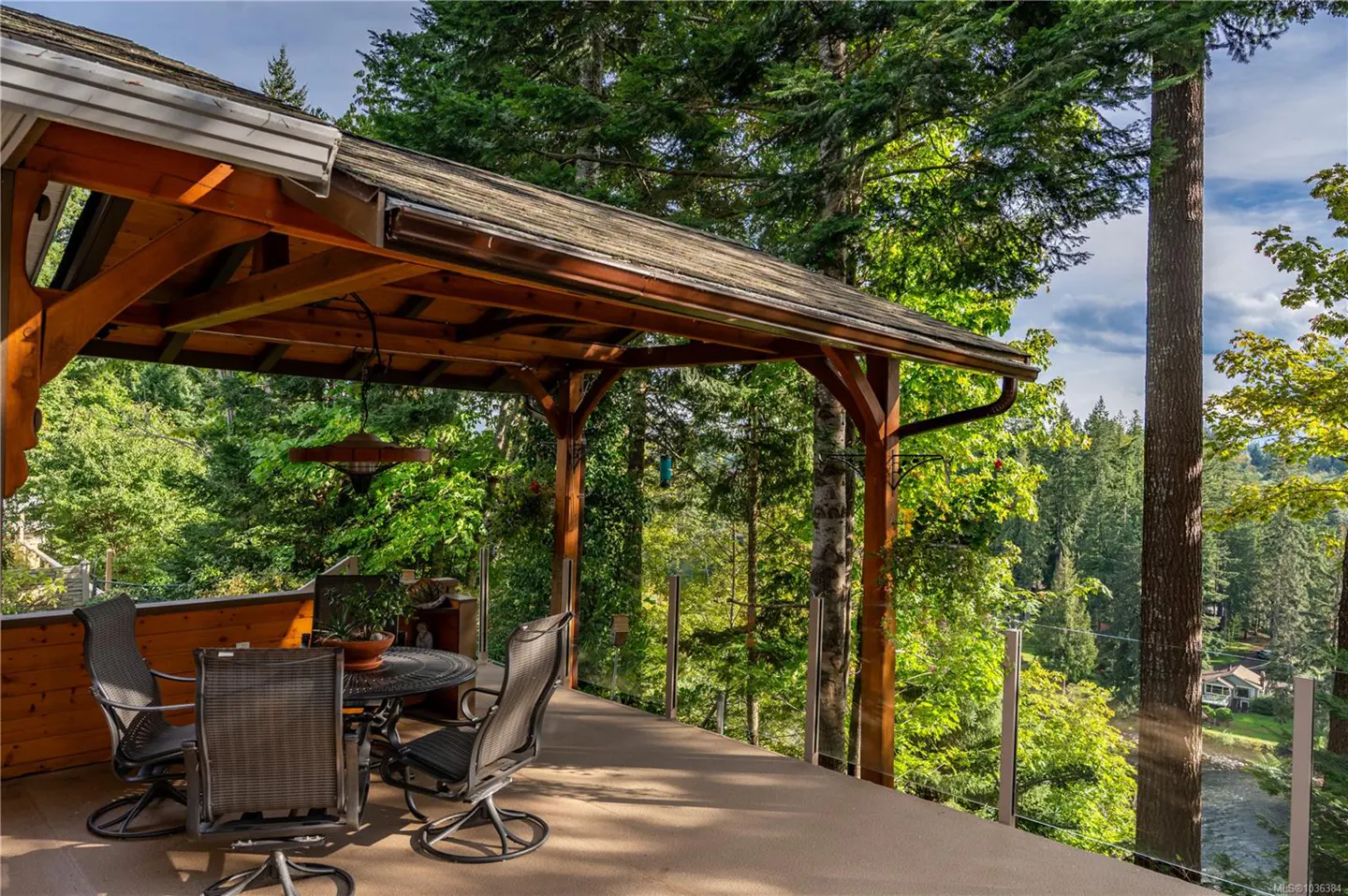 Covered deck with a table and chairs overlooks a lush green forest. The deck has a wooden roof and glass railings.