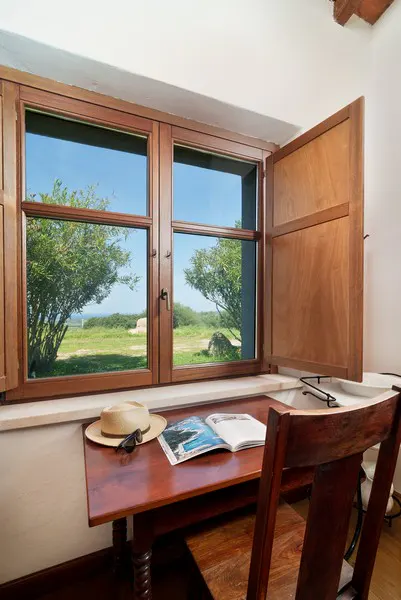 Wooden desk with hat, sunglasses, and open book in front of a window with a view of trees and blue sky.
