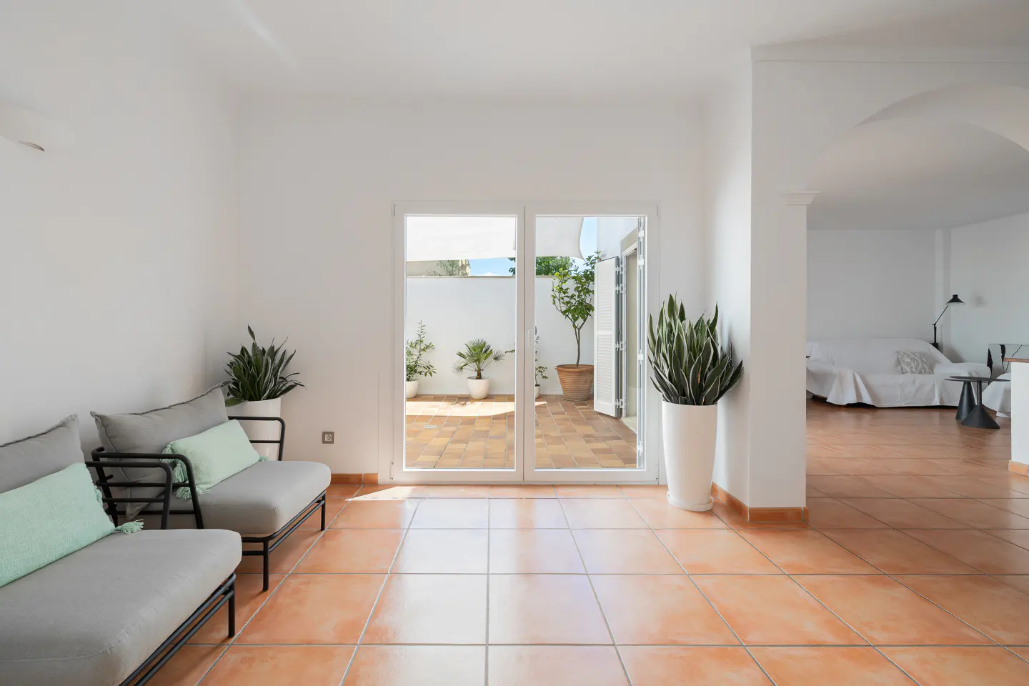Bright, modern living room with terracotta tile floor, white walls, and patio access. Gray chairs with mint pillows sit near potted plants.