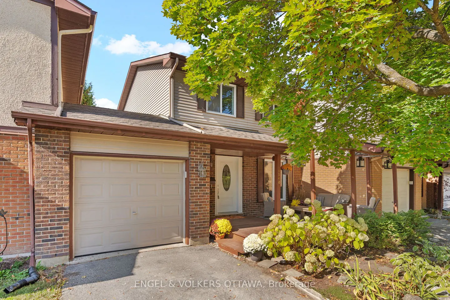 Exterior of a two-story townhouse with a brick facade, beige siding, and a one-car garage. A tree with green leaves is on the right.