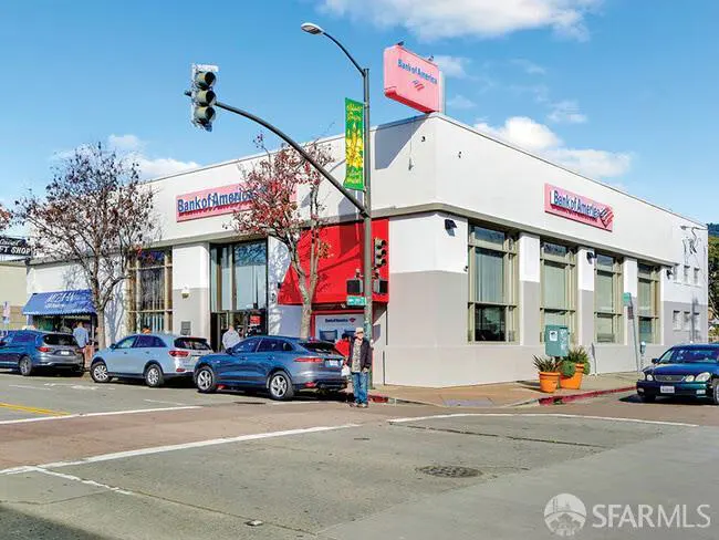 Street view of a Bank of America building with cars parked along the side and a pedestrian crossing the street.