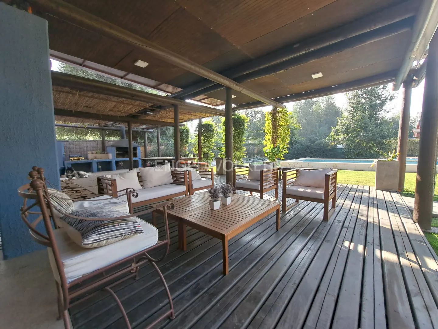 Outdoor patio with wooden deck, sofas, chairs, and table under a covered structure. A pool and greenery are visible in the background.
