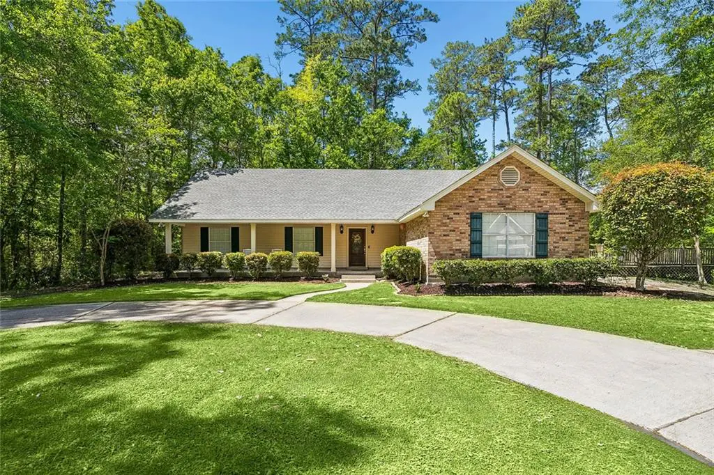 A single-story home with a brick and tan exterior, green shutters, and a gray roof. A curved driveway leads to the front door. Trees surround the property.