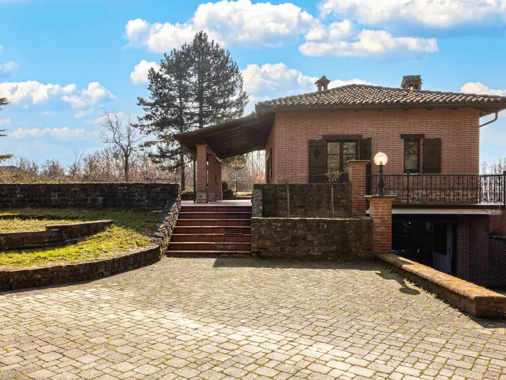 Brick house with a covered porch, stairs, and a stone-paved driveway under a blue sky with white clouds.