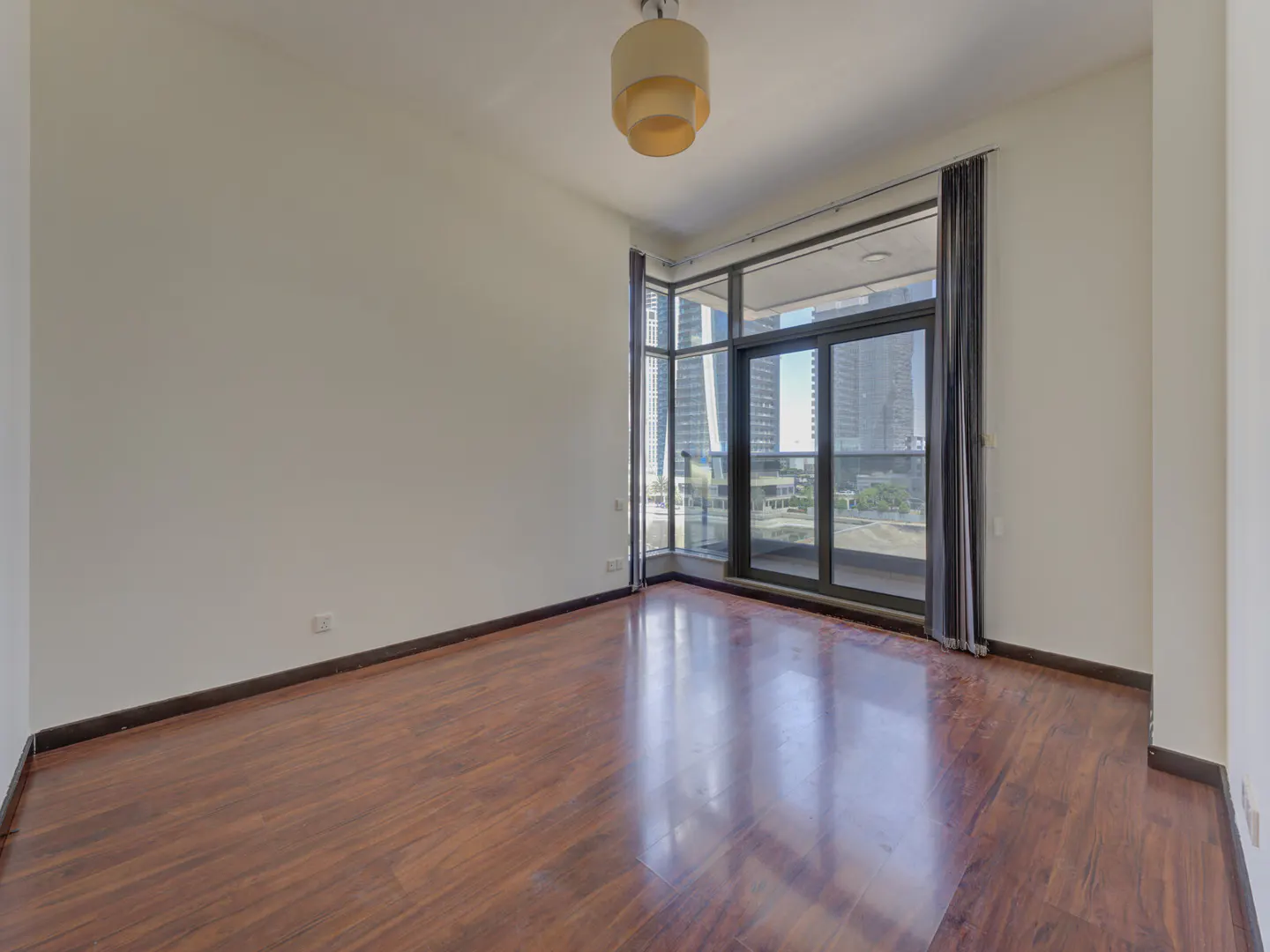 Empty room with hardwood floors, white walls, and a large window with city views. A beige light fixture hangs from the ceiling.