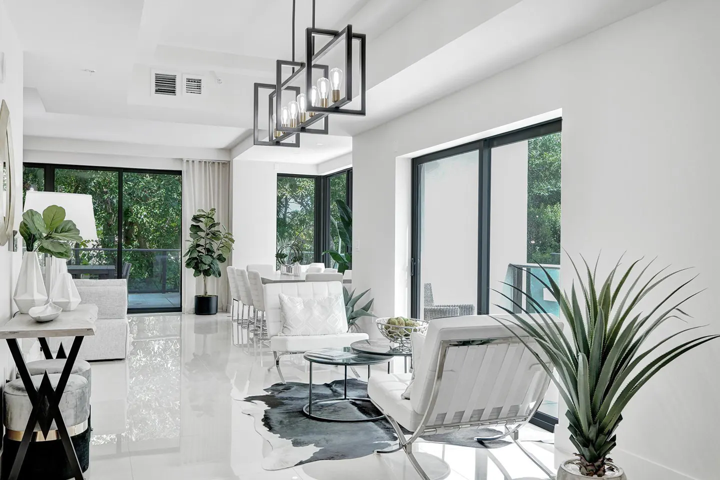 Bright, modern living room with white walls, glossy floors, and large windows. Features white furniture, plants, and a black geometric chandelier.