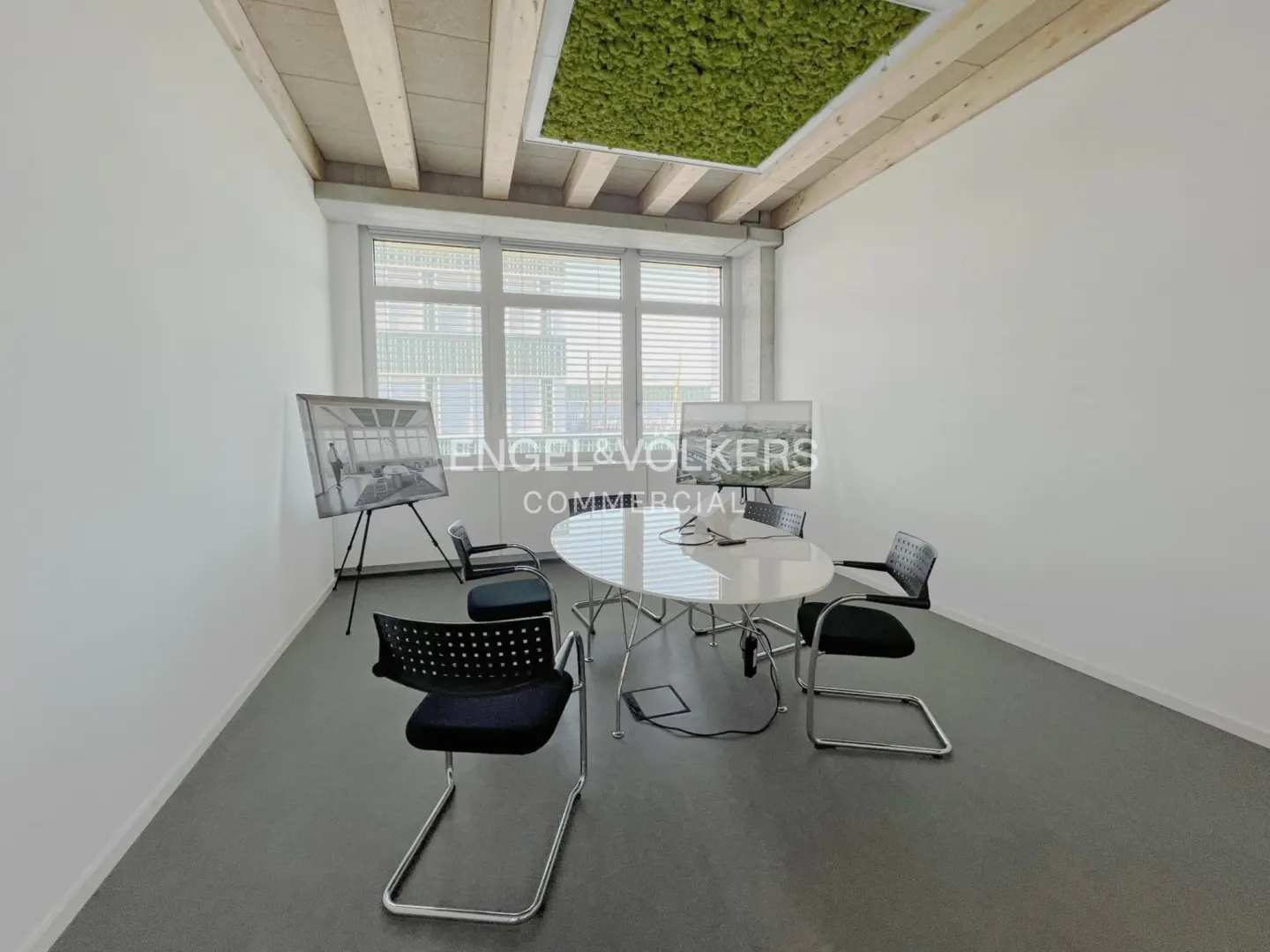 Bright office space with a white table, black chairs, and a green moss ceiling feature. Large windows provide natural light.