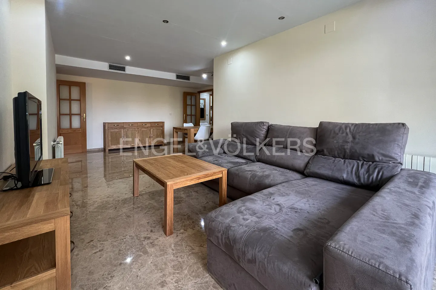Living room with a gray sectional sofa, wood coffee table, TV stand, and marble floors. A dining area is visible in the background.