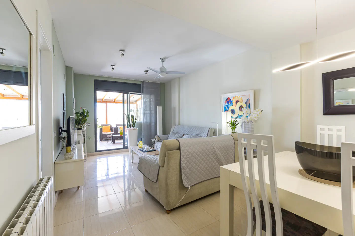 Bright living room with beige sofas, white dining set, and a sliding glass door to a balcony with outdoor furniture.
