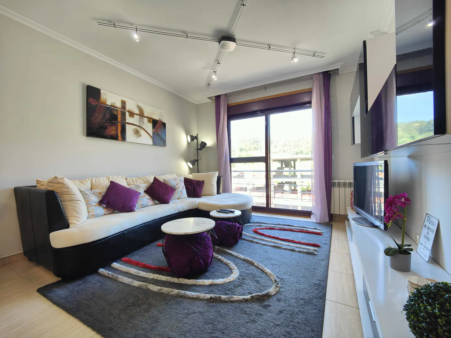 Living room with a black and white sectional sofa, purple pillows, and a gray rug with red and white accents. A TV and balcony are visible.