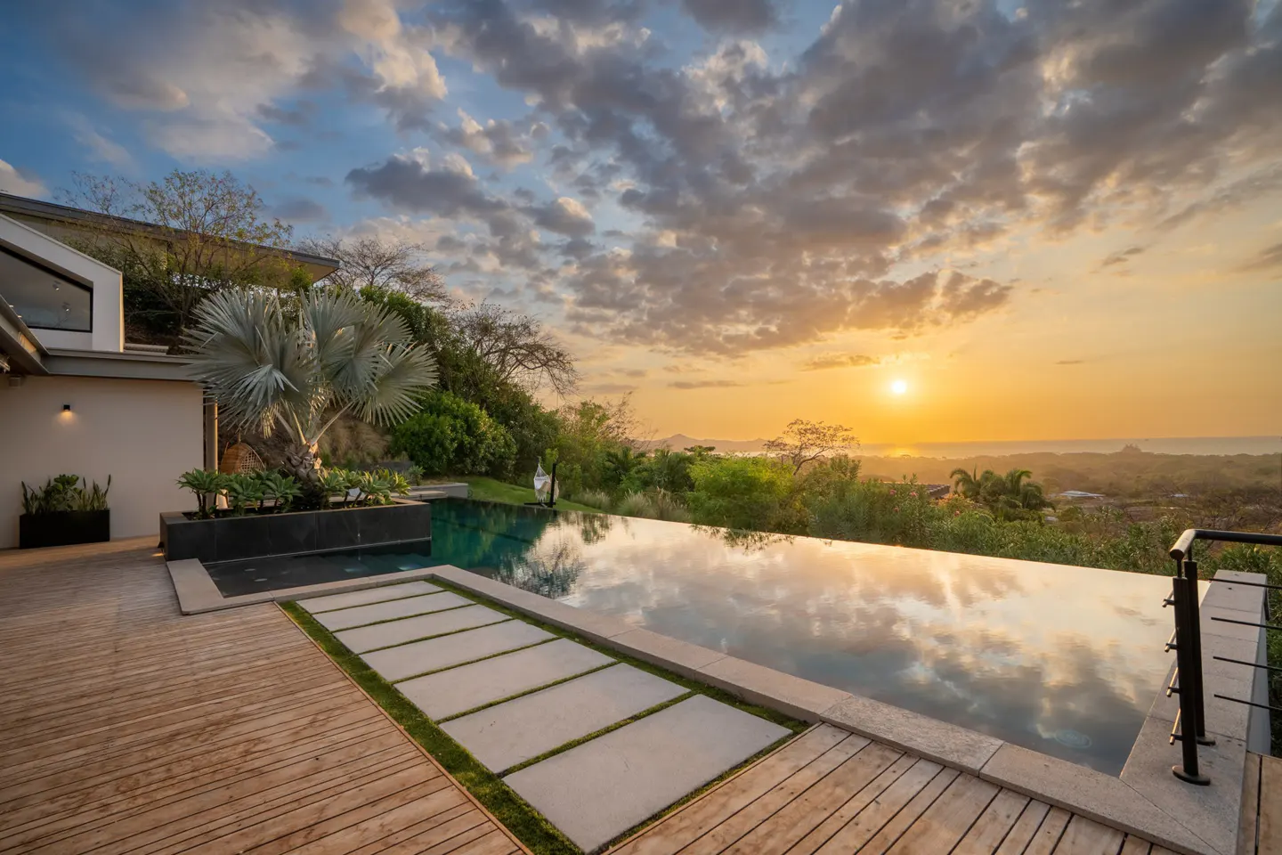 Infinity pool reflecting a sunset sky. Wooden deck and stone pavers lead to the pool's edge, overlooking lush greenery and distant ocean views.