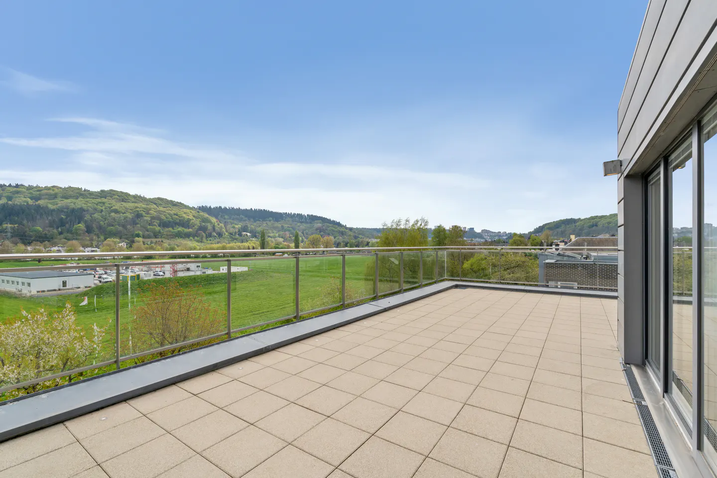 A tiled balcony with a glass railing overlooks a green field and wooded hills under a blue sky.