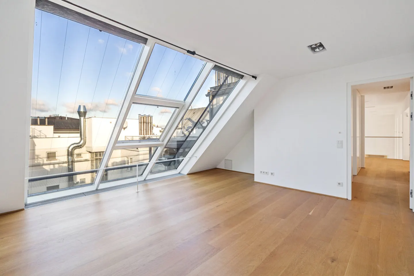 Bright, empty room with wood floors, white walls, and a large angled window showing a cityscape. A doorway leads to another room.